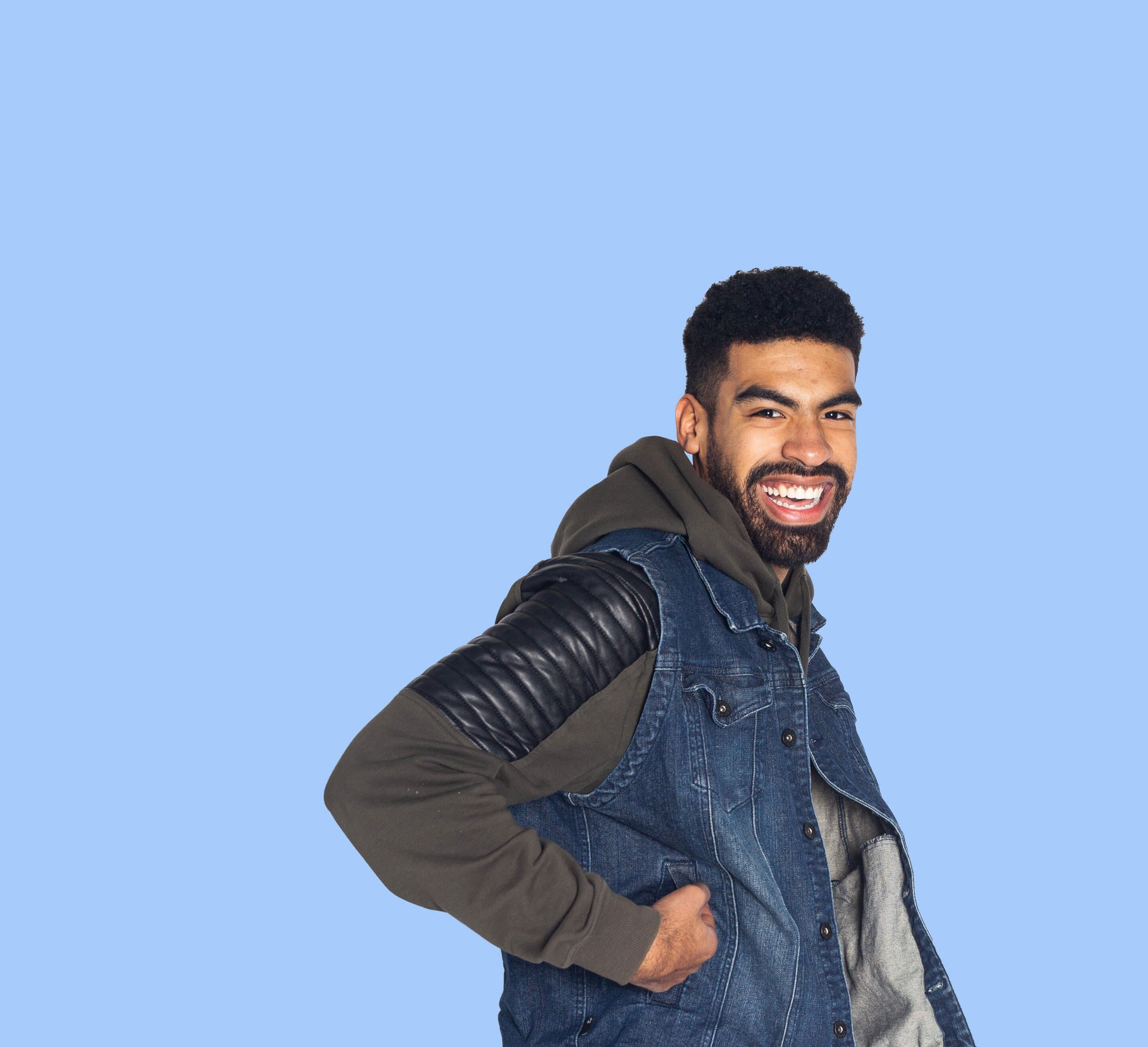 A young man smiling against a blue background wearing a hoodie and denim jacket
