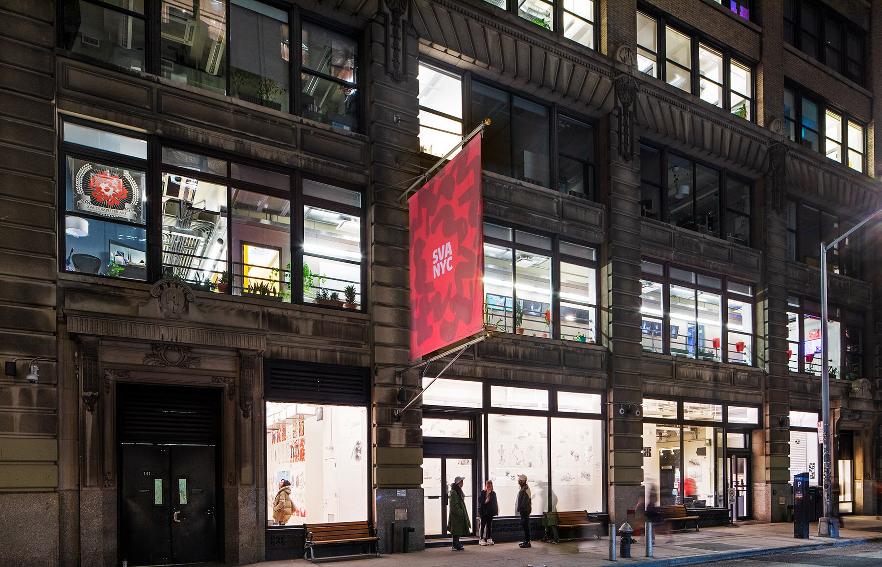 Red SVA Flag on exterior of SVA building at 133 W 21st St, New York, NY at night with students, artwork, and plants visible through the windows